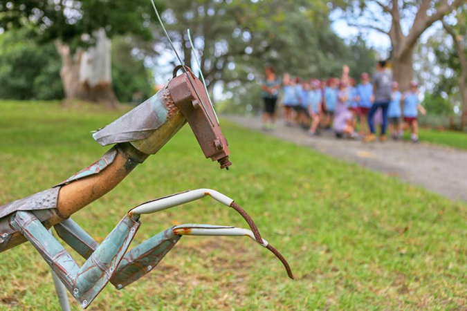 Detail of Libby Bloxham's sculpture with a praying mantis made from found metal on grass, with group of school children out of focus in the background